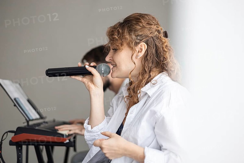 A young Caucasian woman with curly hair sings into a microphone. A man plays a keyboard in the background. The scene captures a home concert atmosphere