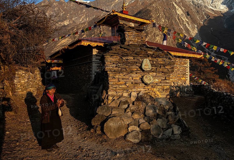 Young woman next to a small Buddhist monastery with manistones in the evening light, Nepalese, local of the Tsumba Tibetan ethnic group traditionally spinning wool with a spindle, Tsum Valley, Himalayas, Nepal
