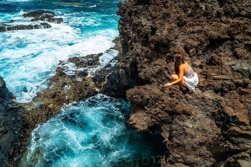 Woman crouching on rugged volcanic rock formations by the ocean, watching turquoise waves crashing and swirling around the unique coastal landscape of el hierro in the canary islands