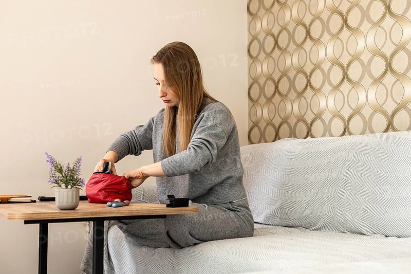 A young Caucasian woman with long brown hair sits at a table in a cozy home setting, unpacking a red makeup bag. A small plant is on the table