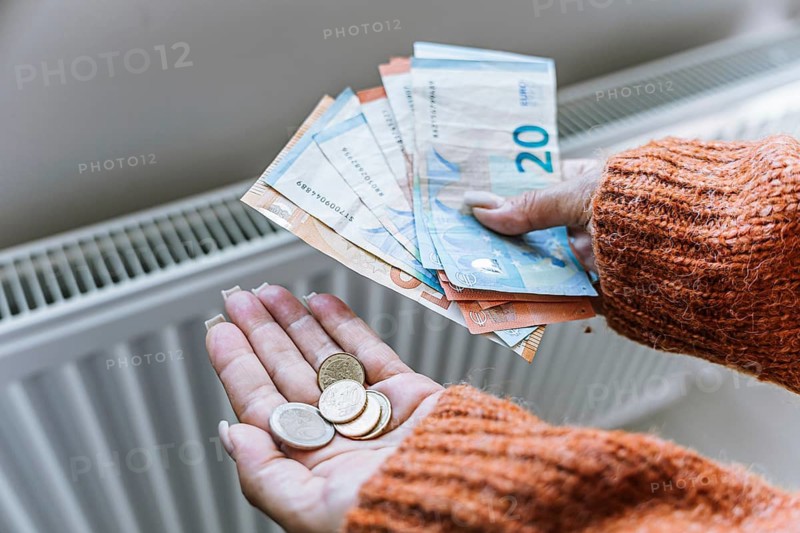 Woman holding euro cash and coins near home radiator during heating season. Concept of rising household energy costs, expensive central heating, utility bills, inflation, cost of living crisis and fin.