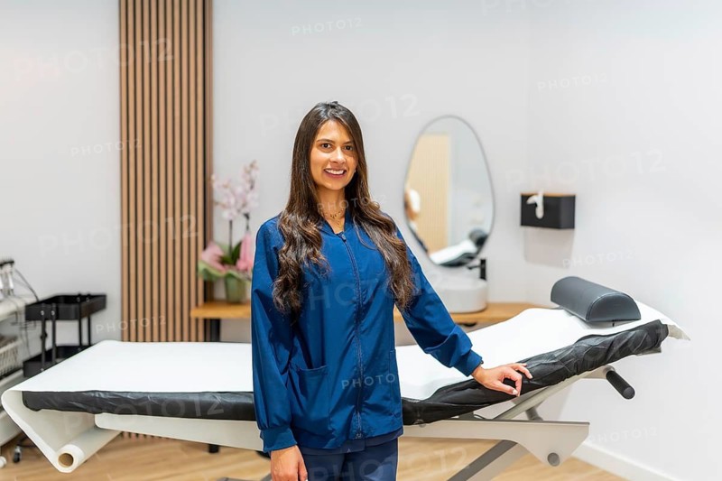 Smiling female professional in blue scrubs standing confidently in a modern aesthetic clinic or laser hair removal center, presenting the clean and equipped treatment room