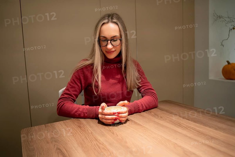 Young woman in red and burgundy sweater with coffee at wooden table, A woman wearing glasses and a red sweater sits at a wooden table holding a cup