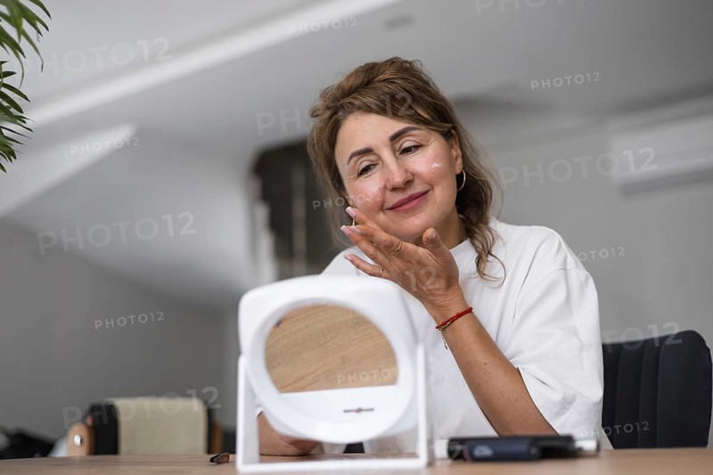 A woman smiles as she applies a skincare product to her face in a well-lit indoor space. The natural light enhances her joyful expression while she interacts with a mirror. Morning routine