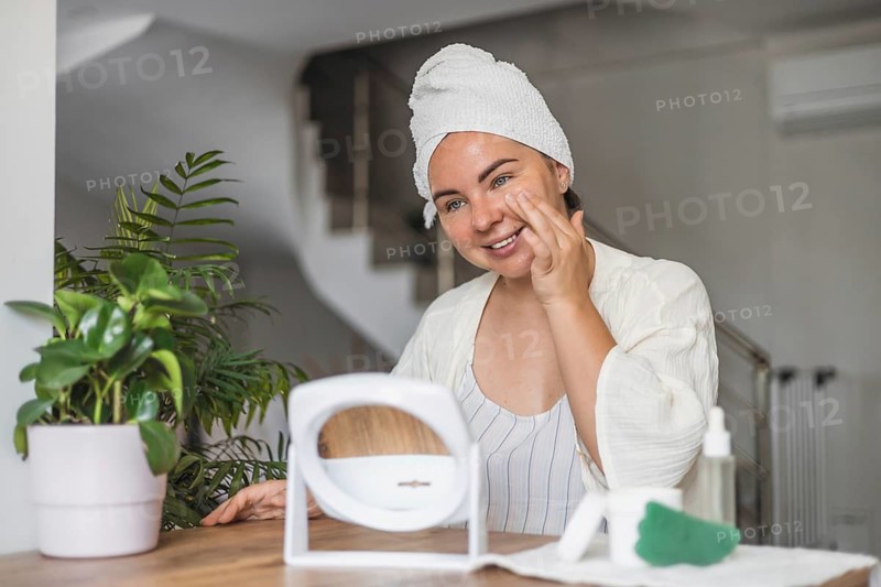 A young Caucasian woman with brown hair in a towel smiles while applying gua sha on her face. A potted plant and beauty products are on the table