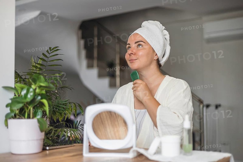 A young Caucasian woman with a towel on her head performs gua sha face massage in a bright room. She holds a jade tool and looks relaxed. A plant and mirror are nearby