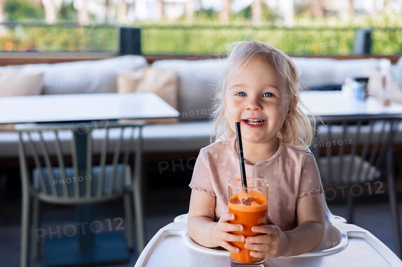 Happy smiling girl toddler drinking carrot juice with a straw in cafe