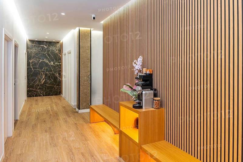 Spacious and inviting hallway in a contemporary aesthetic clinic or wellness center, featuring wooden slat walls, warm lighting, marble textures, and a coffee station, creating a relaxing ambiance