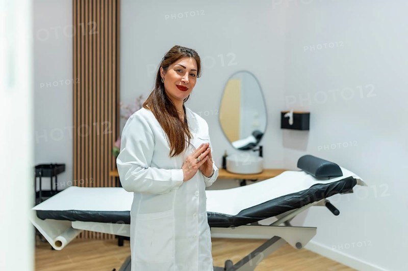 Female beautician or cosmetologist wearing white uniform standing with hands clasped in a clean, contemporary aesthetic clinic and wellness salon, ready for beauty treatments and skin care