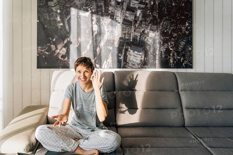 The joyful gesture of a smiling woman who understood how to solve the problem, A woman sitting casually on a couch in a living room, with sunlight and shadow play on a cityscape artwork behind her