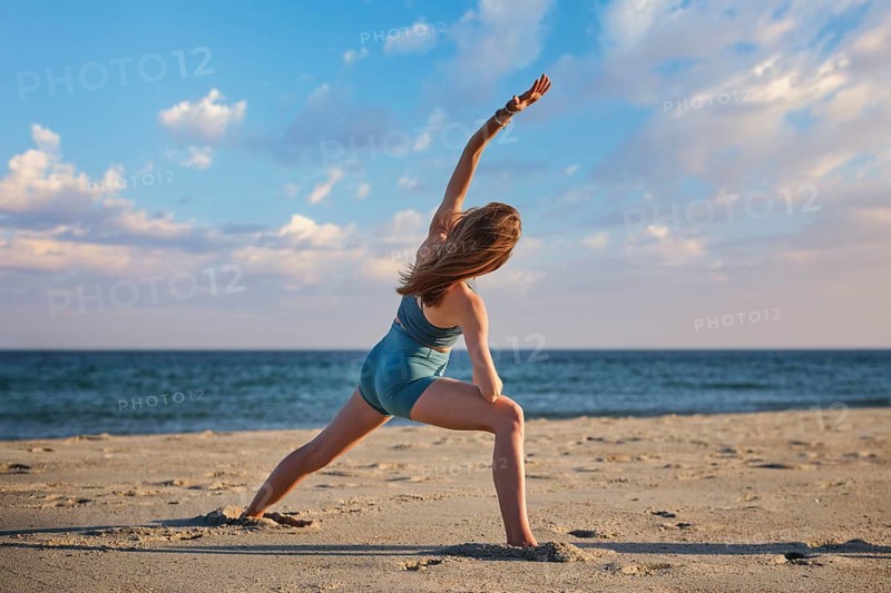 Back view of a fit young woman practicing the utthita parsvakonasana or side angle yoga pose on a beautiful sandy beach by the ocean under a blue cloudy sky, featuring side stretch and stretching