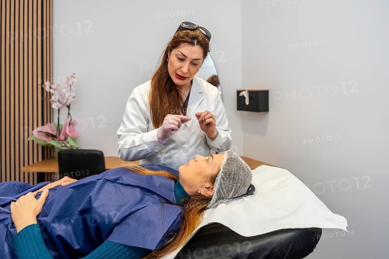 Doctor engaging with a lying patient wearing a hair net and a protective gown, discussing a beauty treatment or facial procedure in a modern aesthetic medicine environment