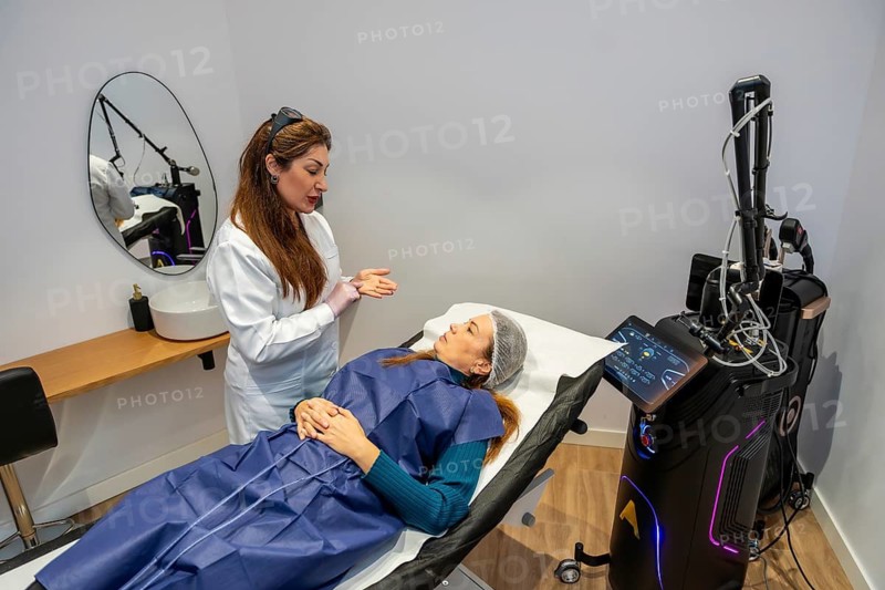 Specialist in protective eyewear explaining laser hair removal procedure to a female client lying on a bed in a modern aesthetic clinic, with advanced cosmetic equipment nearby