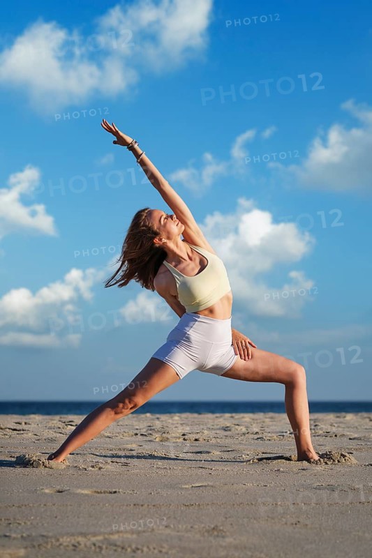 In a half-bound Viparita Virabhadrasana yoga variation, Reverse Warrior, a woman reaches one arm overhead and wraps the other behind her back on beach sand