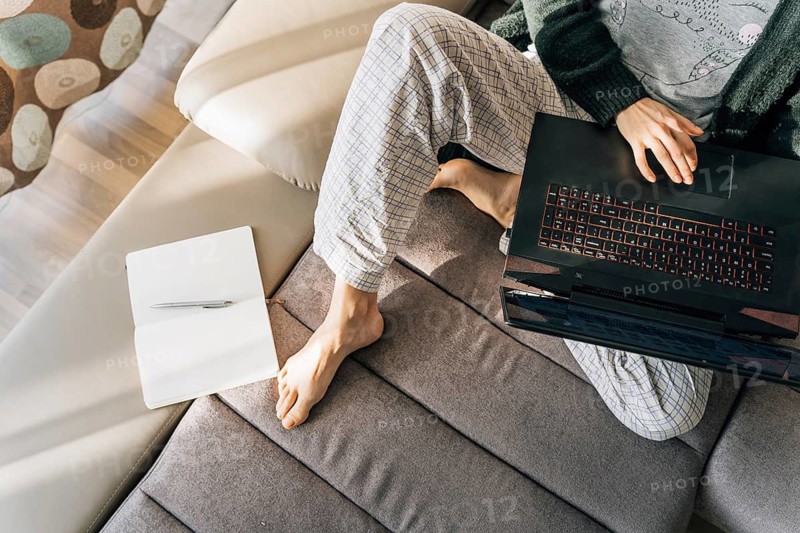 Top view of a woman in pajamas with laptop and notepad, Person sitting on a couch with a laptop and notebook, creating a casual, relaxed work-from-home atmosphere