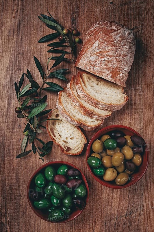 A mixture of olives, varieties of Halkidiki, Verdi giganti and Kalamata, in a bowl, with sliced ciabata bread, top view, no people