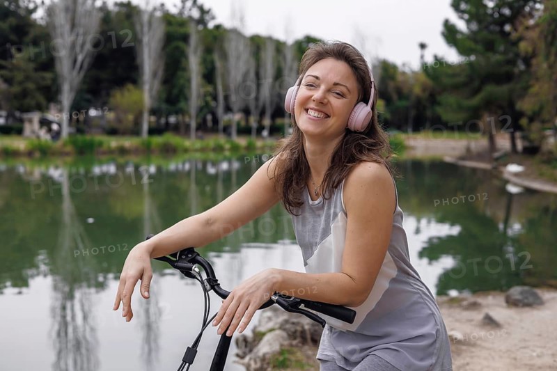 Young Caucasian woman with brown hair and pink headphones smiles while leaning on an electric scooter by a serene lake in a park