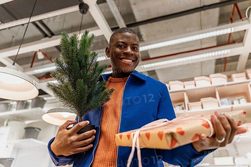 A person stands in a store holding a small potted tree in one hand and a wrapped gift in the other. Shelves with items are visible in the background