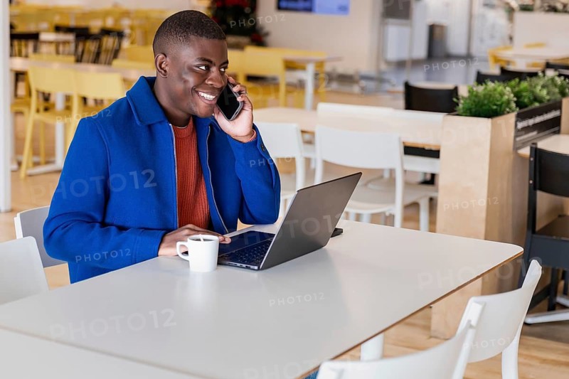 A young African man with short hair is sitting at a table in a modern cafe. He is using a laptop and talking on a smartphone. A cup of coffee is on the table