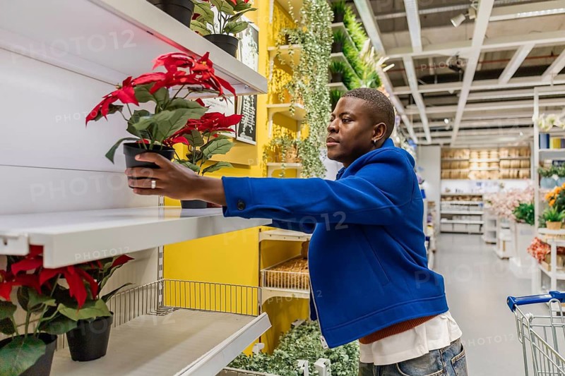 A person stands in a home goods store during the afternoon and arranges a red flower pot on a shelf. The store is filled with various plants and decorations