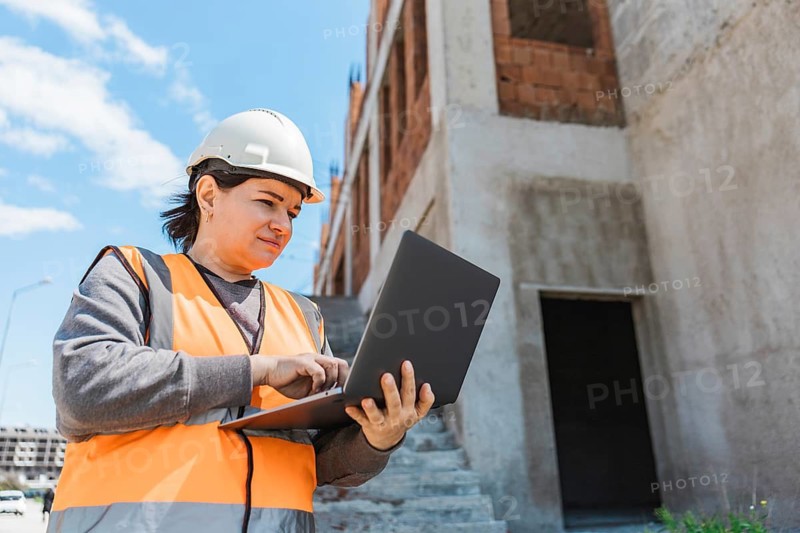 Woman in a hard hat and safety vest uses a laptop at a construction site. The building is in progress with exposed brick and concrete