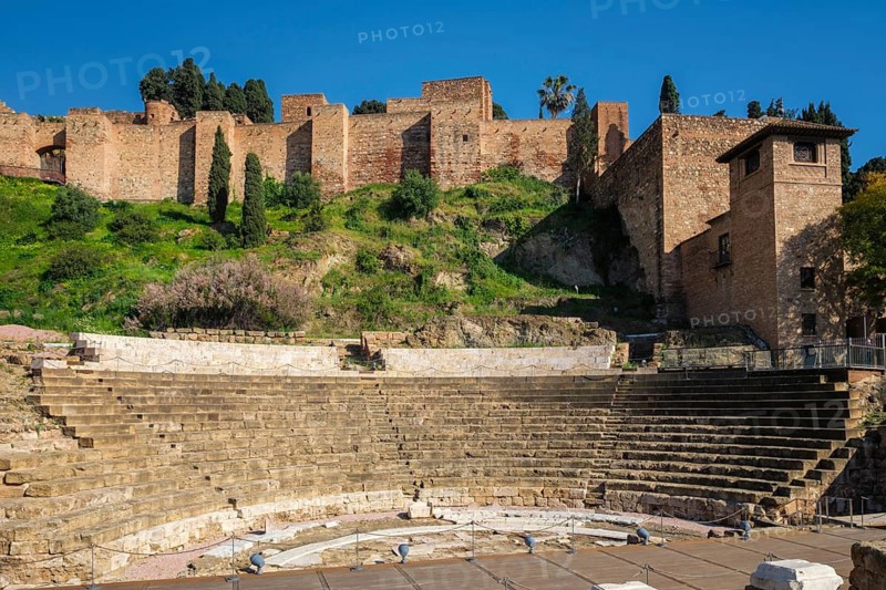 The ancient Roman Theatre ruins in Malaga, Andalusia, Spain, with the historic walls of the Alcazaba Moorish fortress on the hillside above under a clear blue sky, featuring alcazaba of malaga