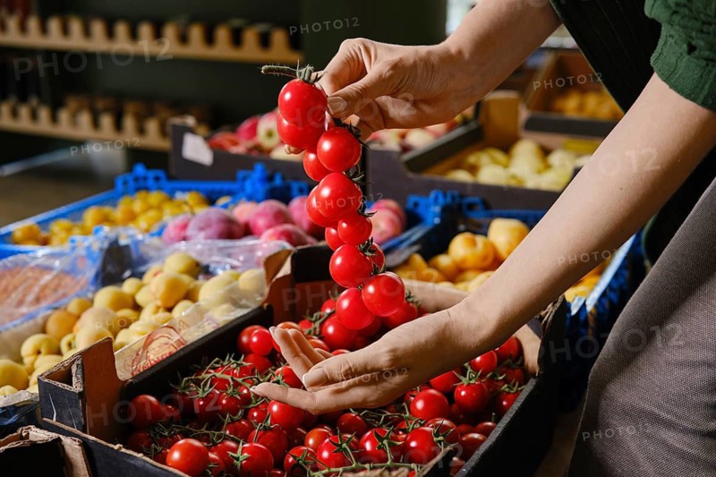 Person holds bunch of red cherry tomatoes in her hand while standing at grocery market. Stalls filled with various fruits like peaches and plums are visible in the background