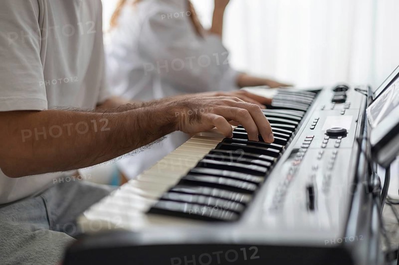 A close-up of a man's hand playing a keyboard in a cozy home setting. A woman is seated nearby, creating a warm atmosphere for a family concert