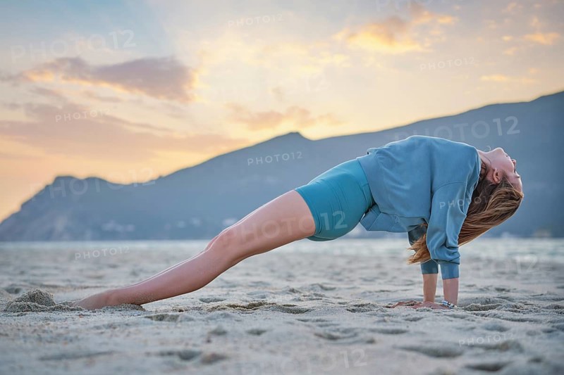 In Purvottanasana yoga, Upward Plank Pose, a woman lifts her hips and chest with straight arms and legs on beach sand beside waves and a coastal hill
