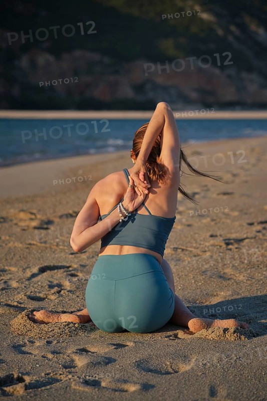 Rear view of a woman practicing a Gomukhasana yoga arm variation, Cow Face Pose, with hands clasped behind her back while seated on beach sand