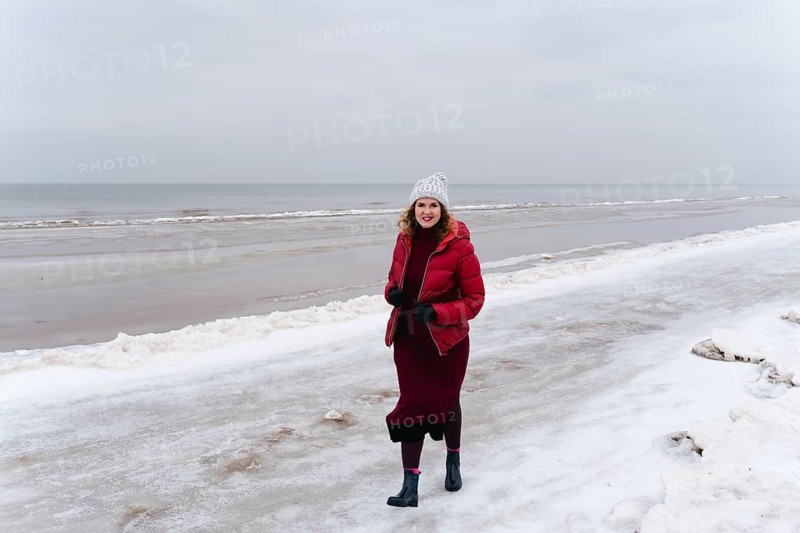 Winter walk along the sea, a woman walks and smiles, A woman in a red coat and hat walks on a snowy beach beside the ocean on a cold, overcast day, Jurmala, Latvia