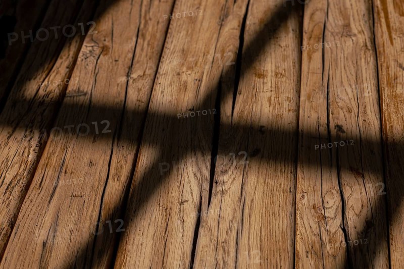 Soft, warm light filters through a window, creating an inviting atmosphere in the room. The light forms distinct shadows on the textured surface of a rustic wooden table, showcasing its knots and grains. This tranquil setting reflects a harmonious blend of nature and home, perfect for moments of relaxation or creativity