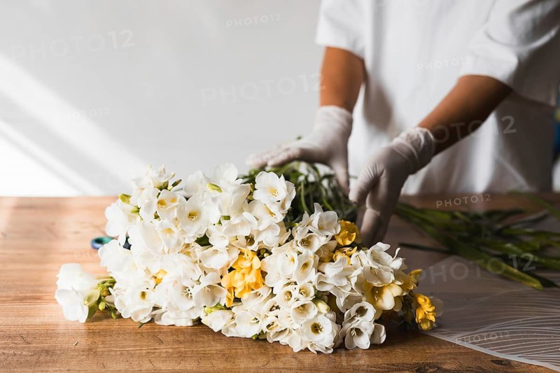 A florist in gloves carefully wraps a bouquet of white flowers with yellow accents on a wooden table. The studio is well-lit and has a calm atmosphere, emphasizing the floral arrangement