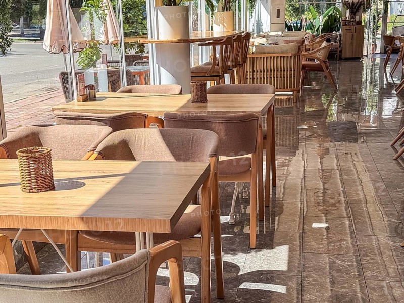 A modern restaurant interior with wooden tables and chairs. The space is bright and airy, featuring large windows and plants for decoration