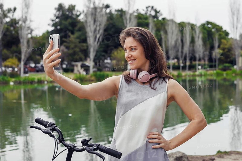 A young Hispanic woman with long brown hair smiles while wearing headphones. She holds a smartphone near a tranquil pond surrounded by greenery