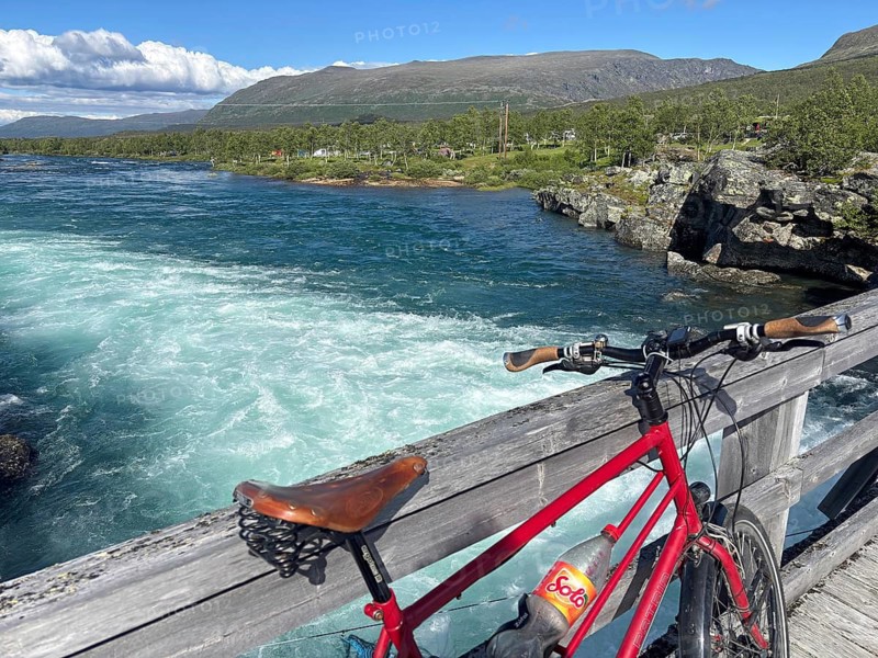 Red bicycle standing on a wooden bridge overlooking a rushing river and mountains, Sjoa, Gjendersheim, Maurvangen, Jotunheimen, Norway