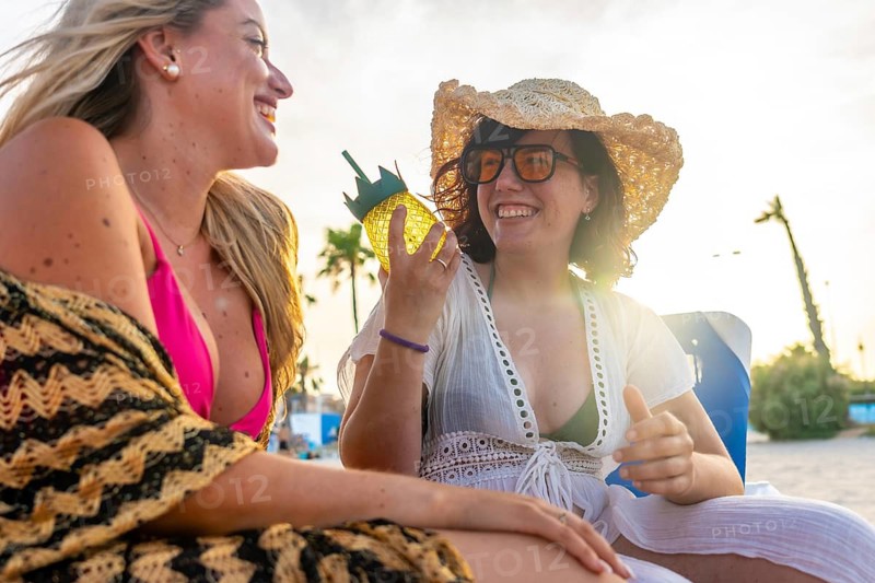 Two young women laughing and relaxing on a beach lounge chair at golden hour, celebrating friendship and enjoying a tropical drink together during a summer holiday