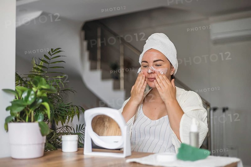 A woman in a towel applies cream to her face while seated at a wooden table