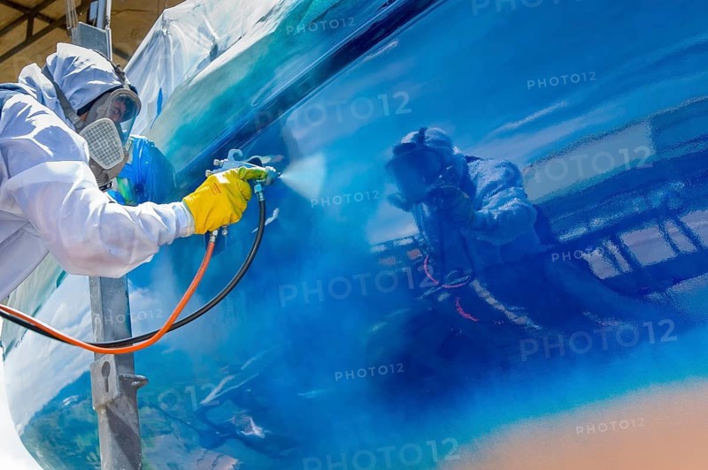Worker spray paints the hull of a sailing yacht during professional marine maintenance, wearing protective clothing and equipment