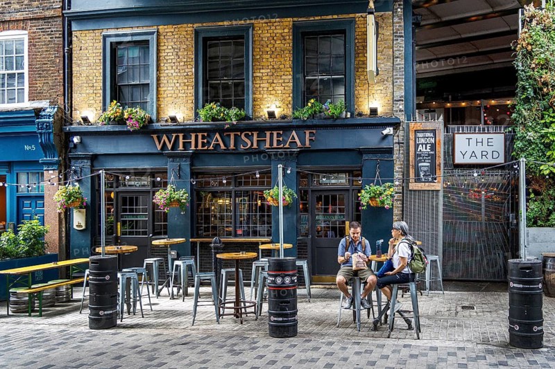 Two visitors sit at outdoor tables made from upturned beer casks outside the Wheatsheaf, a traditional Southwark pub with black timbered windows and overflowing hanging baskets, next to the entrance to The Yard near Borough Market in London, England, United Kingdom