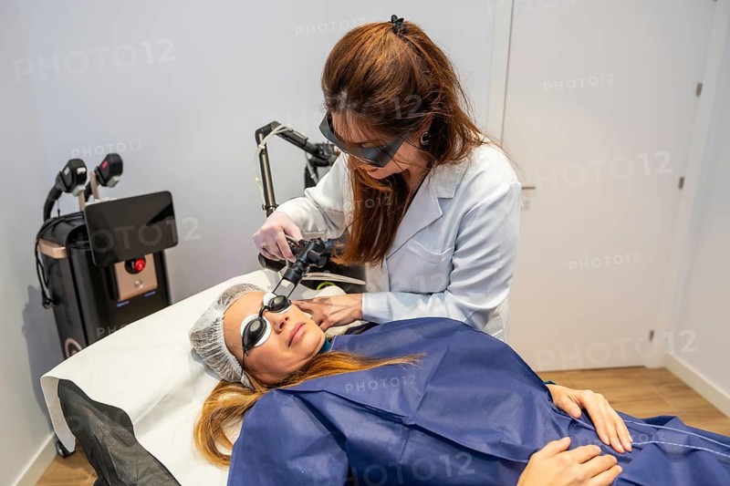 Medical professional in protective eyewear and a lab coat applying a laser device to a female patient's face for a skin treatment in a modern aesthetic clinic
