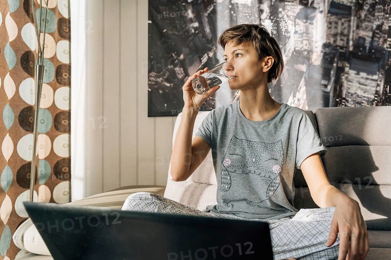 Young blonde drinks water from a glass sitting at home with a laptop, A woman in casual clothing drinking water while sitting on a couch with sunlight streaming in