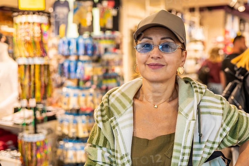 Middle-aged Caucasian woman with glasses and a cap stands in a souvenir shop. Colorful merchandise and displays are visible in the background