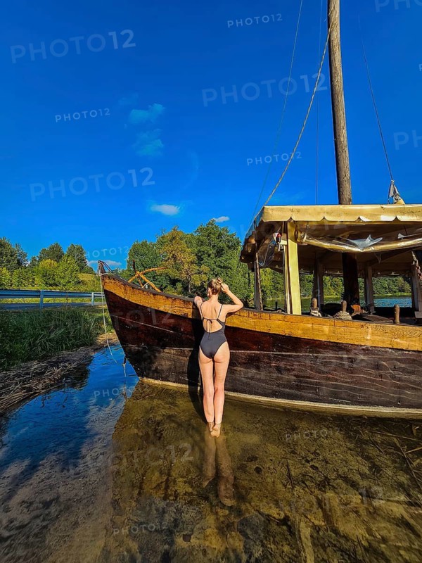 Rear view of a slim fit woman in a black one-piece swimsuit standing in a lake near a wooden vintage ship, A woman in swimwear stands near a wooden boat on a sunny day with a clear blue sky
