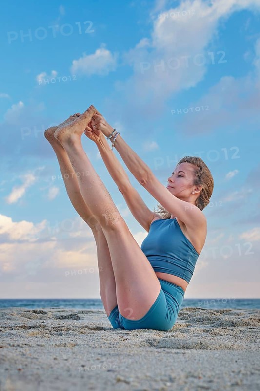 A woman practices Ubhaya Padangusthasana yoga, Both Big Toe Pose, in a seated balance on beach sand, holding both raised feet beneath a blue sky