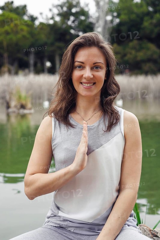 A young Caucasian woman with brown hair sits by a serene lake, practicing yoga. She smiles and holds a hand gesture, surrounded by greenery and water