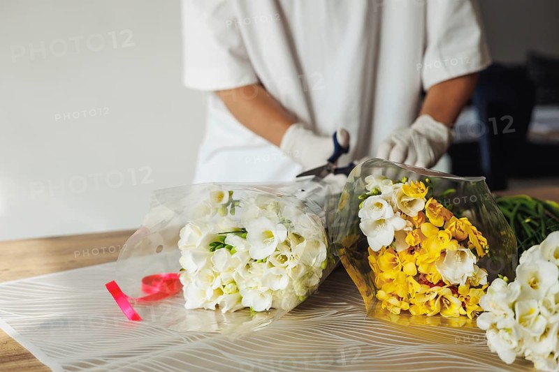 A florist, wearing gloves, is carefully arranging white and yellow flowers in wrapping paper. Scissors and ribbons are visible in the background, highlighting the vibrant workspace and creativity