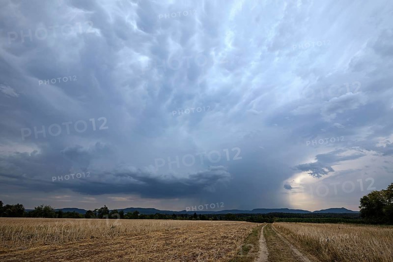 Dark storm clouds and heavy rain gather over a harvested field in Germany, with a dirt road leading toward the Swabian Alb hills