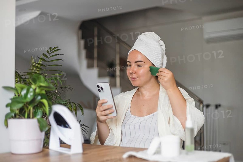 A young Caucasian woman with brown hair in a towel uses a green gua sha tool for facial massage while looking at her phone in a bright room with plants