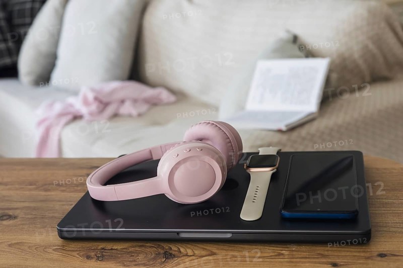 A cozy living room scene with a young woman reading a book. On a wooden table, there are pink headphones, a smartwatch, and a smartphone